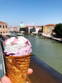 Cropped image of hand holding ice cream cone by canal against sky
