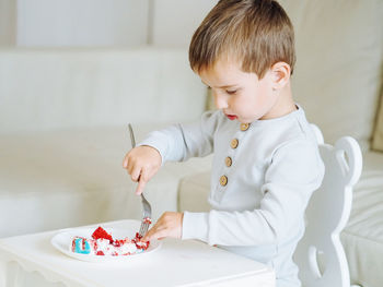 Cute toddler 3 years boy in grey pajamas eating red velvet cake in bright interior