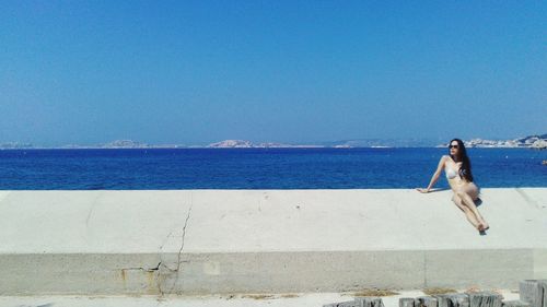 Young woman in bikini on retaining wall by sea against clear sky