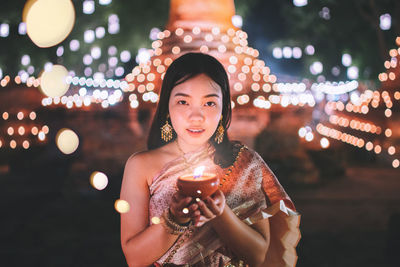 Portrait of young woman holding diya while standing against illuminated temple at night