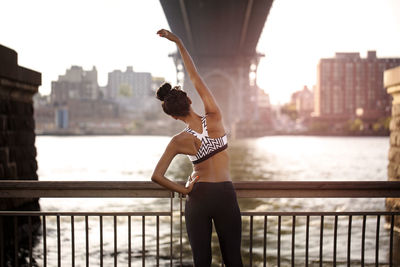 Rear view of sporty woman stretching by railing under williamsburg bridge