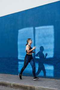 Young woman jogging on footpath by blue wall