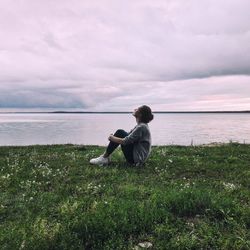 Rear view of man sitting on grass by sea against sky