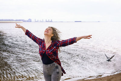 Smiling woman with arms outstretched standing at beach against sky