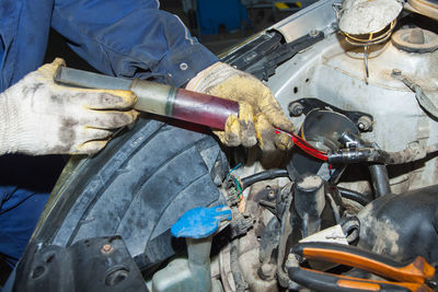 The mechanic's gloved hands hold a syringe of red oil