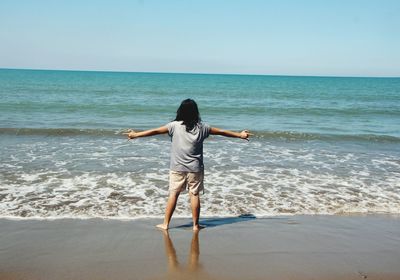 Rear view of woman standing with arms outstretched on shore against sky