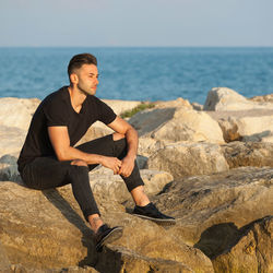 Young man sitting on rock by sea against sky