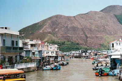 Boats on river amidst buildings and mountains against clear sky