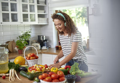 Smiling teenage girl listening music while chopping vegetables in kitchen