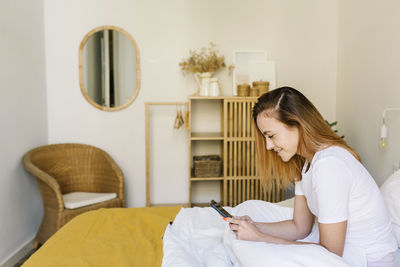 Young woman using phone while sitting on bed at home