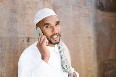 Cheerful muslim male in traditional clothes smiling and browsing cellphone while standing near shabby wall on street