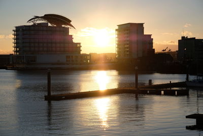 Silhouette buildings by river against sky during sunset