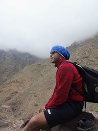 Young man sitting on mountain against sky