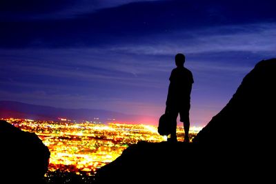 Silhouette man standing on mountain overlooking illuminated cityscape at night