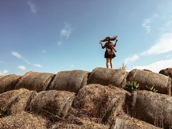 Rear view of woman jumping on hay bales stack against sky