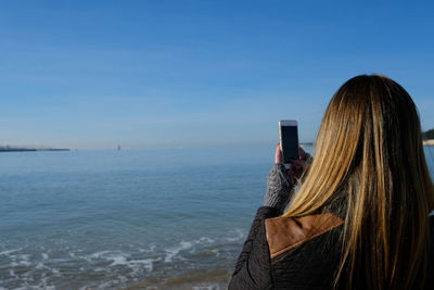 Rear view of woman photographing sea with smart phone against sky