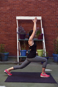 Side view of woman with arms raised practicing warrior 1 pose against brick wall