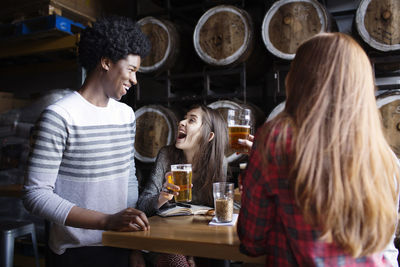 Friends enjoying while having beer at table against barrels in brewery