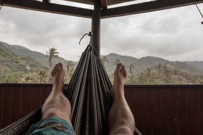 Low section of man resting on hammock in balcony by mountains against sky