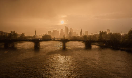 Bridge over river by buildings in city against sky during sunset