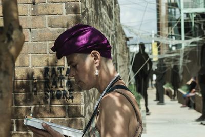 Side view of woman reading book by brick wall in city