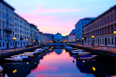 Tilt-shift image of boats moored in canal amidst buildings against church during sunset