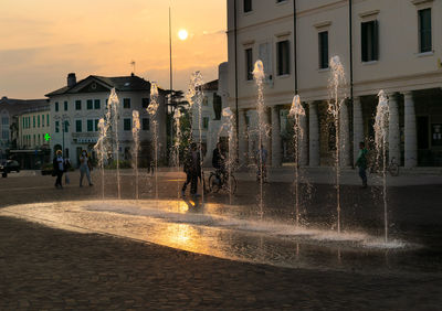 People walking on wet street by buildings against sky during sunset