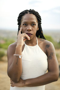 Adult black female in white top with afro braids touching face and looking at camera on blurred background of countryside