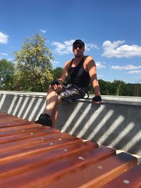 Portrait of man wearing sunglasses sitting on retaining wall against blue sky