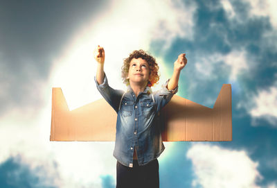 Thoughtful boy with cardboard wings against cloudy sky