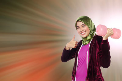 Portrait of smiling young woman gesturing thumbs up while holding dumbbell against abstract backgrounds