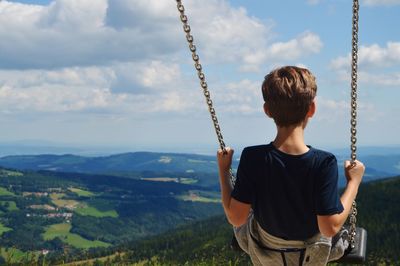 Rear view of boy swinging by landscape against cloudy sky