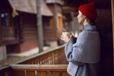 Woman in a red hat and scarf and a mug stands at a wooden house, in the woods in the autumn morning 