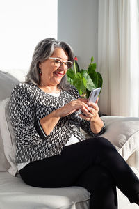 Smiling mature female in classic outfit and eyeglasses browsing modern mobile phone while sitting with legs crossed on comfy sofa