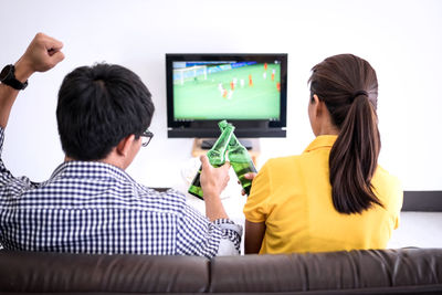 Rear view of friends toasting beer bottles while watching sports on tv at home