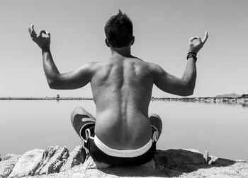 Rear view of shirtless man mediating on rock by sea against clear sky