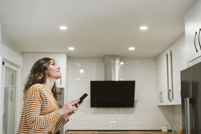 Young woman using mobile phone with smart home app for ceiling lights at home
