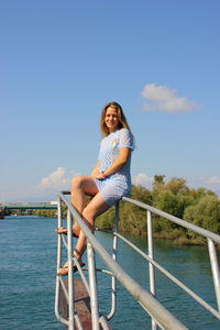 Portrait of young woman sitting on railing over river against sky