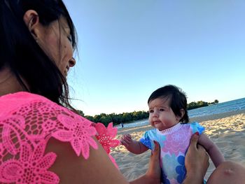 Side view of mother with cute daughter sitting at beach against clear blue sky during sunset