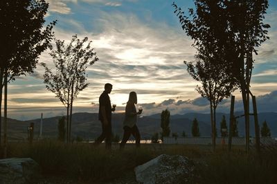 Couple holding wineglass walking on grassy field by mountains against sky during sunset