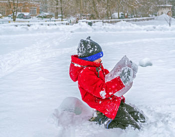 Boy in a red jacket and knitted hat is sitting in the snow and holding a large block of ice.