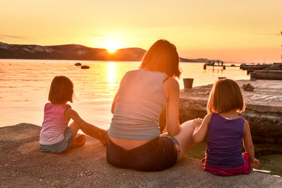 Rear view of mother with daughters resting by sea during sunset