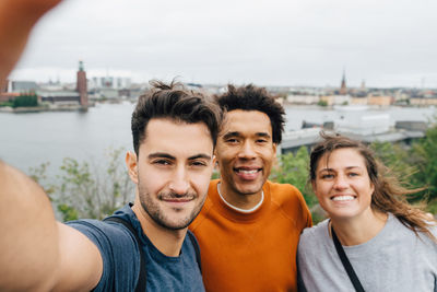 Portrait of smiling friends taking selfie while standing in city against sky