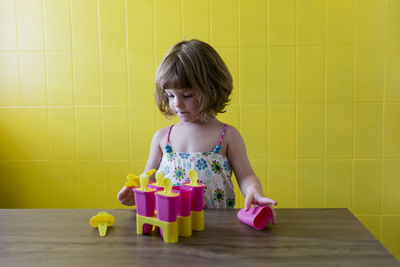 Cute girl playing with toys on table against yellow wall