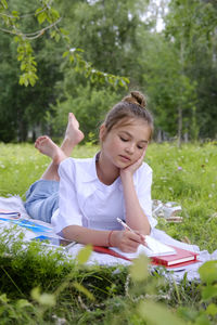 A young schoolgirl lies on the grass in the park with a notebook and a pen.