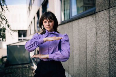 Portrait of young woman gesturing equal sign while standing against building and wall