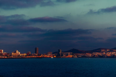 Illuminated buildings by sea against sky at dusk