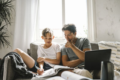 Son looking in book while father sitting by with laptop on sofa at home