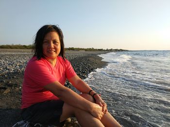 Portrait of smiling young woman sitting on beach against clear sky