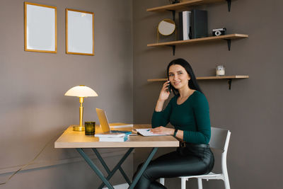 Young caucasian woman is sitting at home at her desk with a laptop and talking on a mobile phone